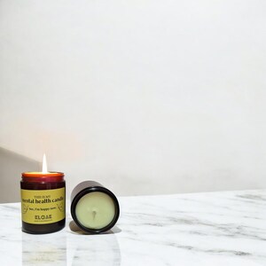 May include: A lit candle in a brown glass jar with a yellow label that reads "THIS IS MY mental health candle See, I'm happy now." The candle is next to an identical jar with the lid off. The background is white.
