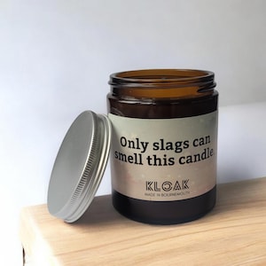 Puede incluir: Un tarro de vela de vidrio marr&oacute;n con tapa plateada, sobre una superficie de madera. La etiqueta dice "Only slags can smell this candle" y la marca "KLOAK".