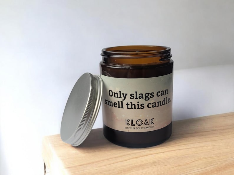 Puede incluir: Un tarro de vela de vidrio marr&oacute;n con tapa plateada, sobre una superficie de madera. La etiqueta dice "Only slags can smell this candle" y la marca "KLOAK".