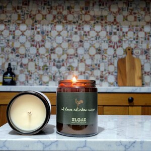 May include: Two lit, brown glass jar candles with white wax and a label that reads "I love chicken wine" and "KLOAK". One candle is open, the other is lit. The background includes a colorful tile backsplash and wooden cabinets.