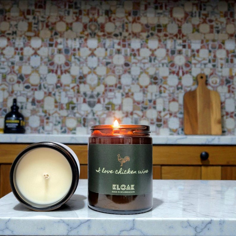 May include: Two lit, brown glass jar candles with white wax and a label that reads "I love chicken wine" and "KLOAK". One candle is open, the other is lit. The background includes a colorful tile backsplash and wooden cabinets.