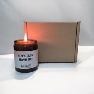 Pode incluir: Uma vela de vidro marrom com um r&oacute;tulo branco que diz "HOT GIRLS HAVE IBS" e "KLOAK MADE IN BOURNEMOUTH". A vela est&aacute; acesa e a chama &eacute; vis&iacute;vel. A vela est&aacute; sentada em uma superf&iacute;cie branca na frente de uma caixa de papel&atilde;o marrom.