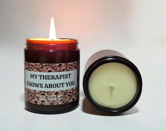 May include: A brown glass candle jar with a black lid. The candle is lit and the jar has a label that reads "My therapist knows about you."