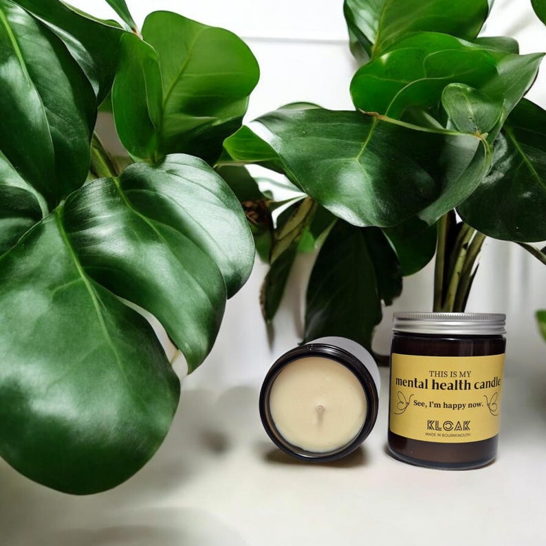 May include: Two candles are displayed in front of a green plant. One is in a jar with a yellow label reading "THIS IS MY mental health candle" and "See, I'm happy now." The other is an open black candle. The candles are on a white surface.