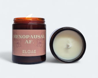 May include: A brown glass jar candle with a lit wick and a matching lid. The label reads "MENOPAUSAL AF." with floral accents and the brand name "KLOAK." The candle is on a white background.