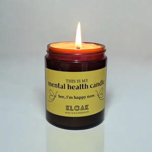 May include: A burning candle in a brown glass jar with a yellow label. The label says "THIS IS MY mental health candle See, I'm happy now." The candle is from KLOAK, made in Bournemouth.