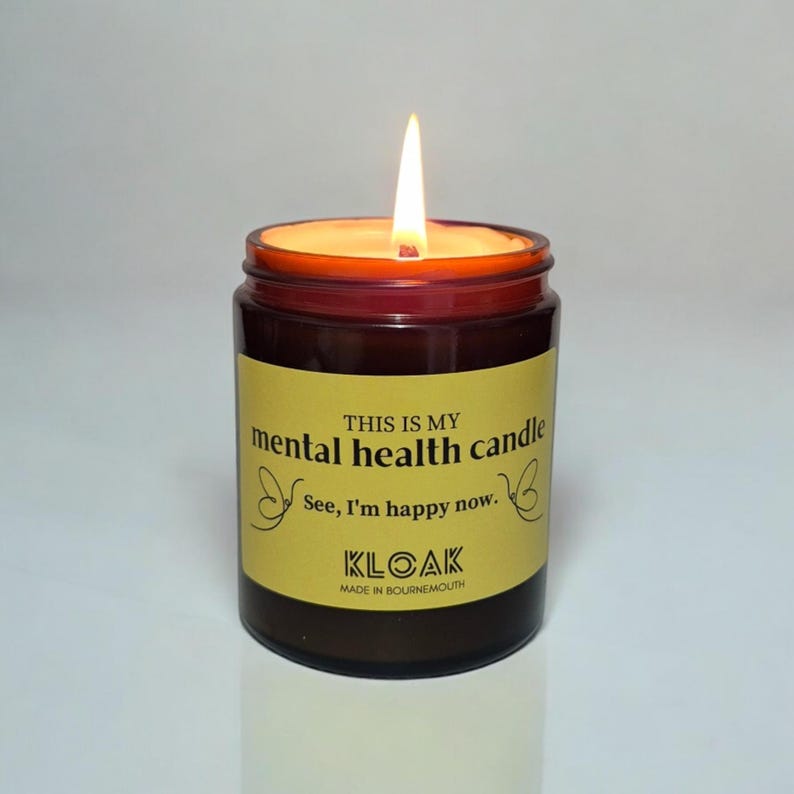 May include: A burning candle in a brown glass jar with a yellow label. The label says "THIS IS MY mental health candle See, I'm happy now." The candle is from KLOAK, made in Bournemouth.