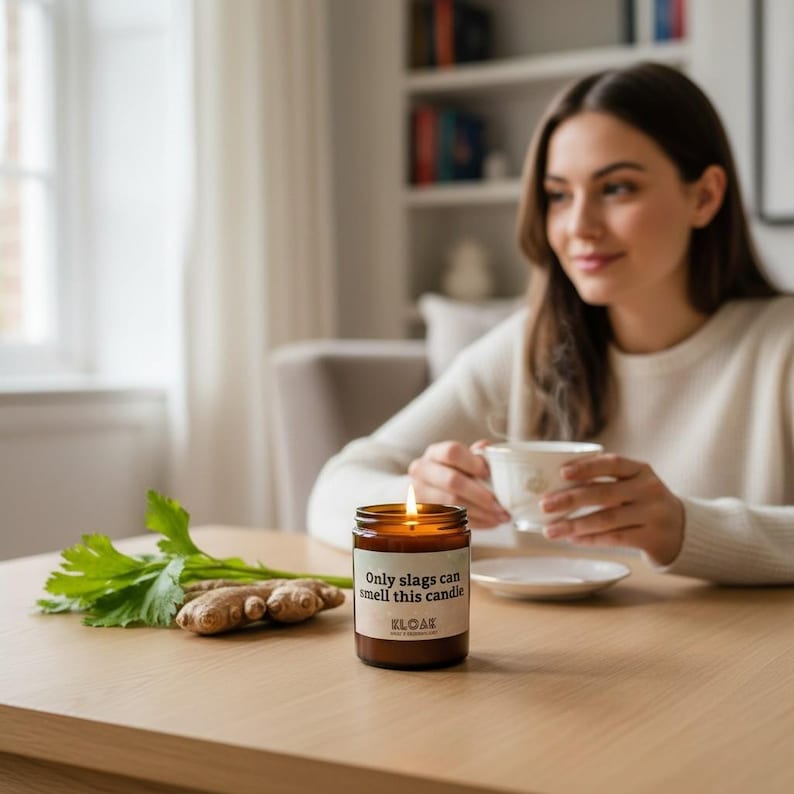 Puede incluir: Una vela encendida en un tarro de cristal marr&oacute;n con el texto "Only slags can smell this candle" y la marca "KLOAK" en una etiqueta. La vela est&aacute; sobre una mesa de madera junto a jengibre y apio. Una persona est&aacute; en el fondo.