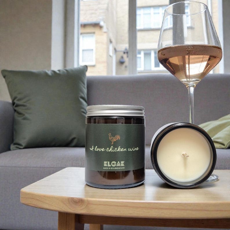 May include: A candle in a glass jar with a silver lid and a label that reads "I love chicken wine" and "KLOAK". A wine glass with a pink liquid is next to the candle. The candle is on a wooden table.