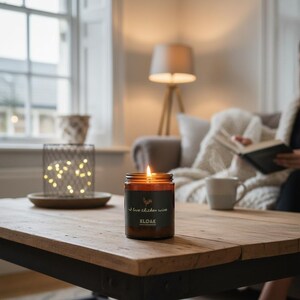 May include: A lit brown glass candle jar with a black label that reads "I love chicken wine" and the brand name "KLOAK". The candle sits on a wooden table. A metal wire basket with fairy lights is in the background.