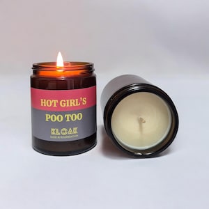 Puede incluir: Un tarro de vela de vidrio marrón con una etiqueta roja y gris que dice "HOT GIRL'S POO TOO" y "KLOAK MADE IN BOURNEMOUTH". La vela está encendida y la llama es visible.