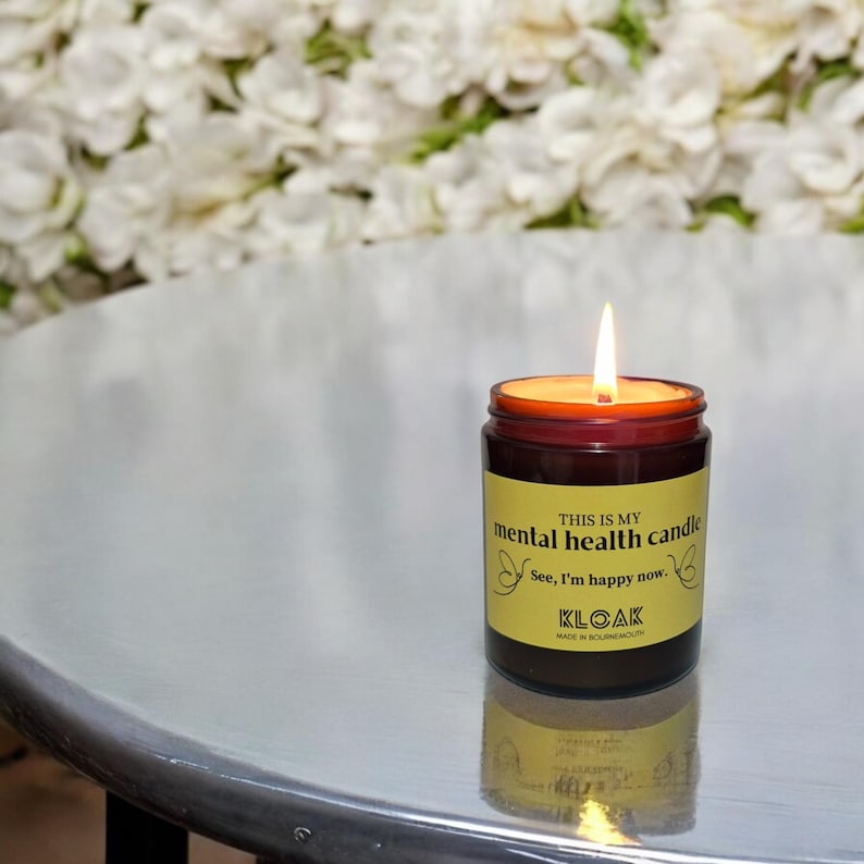 May include: A burning candle in a brown glass jar with a yellow label. The label says "THIS IS MY mental health candle. See, I'm happy now." The candle is on a reflective surface, with a floral backdrop.