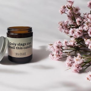 Puede incluir: Un frasco de vela de vidrio marr&oacute;n con tapa plateada y una etiqueta que dice Only slags can smell this candle y KLOAK. La vela est&aacute; colocada junto a un racimo de peque&ntilde;as flores rosas con tallos y hojas verdes.