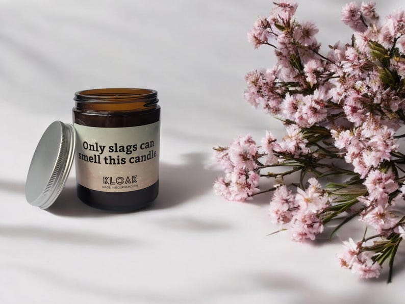 Puede incluir: Un frasco de vela de vidrio marr&oacute;n con tapa plateada y una etiqueta que dice Only slags can smell this candle y KLOAK. La vela est&aacute; colocada junto a un racimo de peque&ntilde;as flores rosas con tallos y hojas verdes.