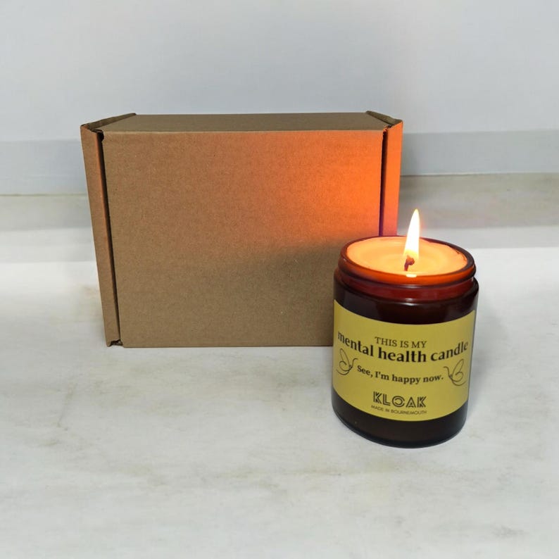 May include: A burning candle in a brown glass jar with a yellow label. The label says "This is my mental health candle. See, I'm happy now." A brown cardboard box is behind the candle. The candle has a bright orange glow.