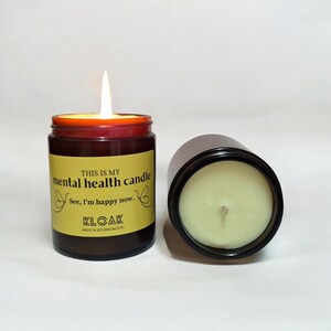 May include: A lit, brown glass jar candle with the text "THIS IS MY mental health candle. See, I'm happy now." and the brand name "KLOAK". A second, unlit candle of the same type is next to it.