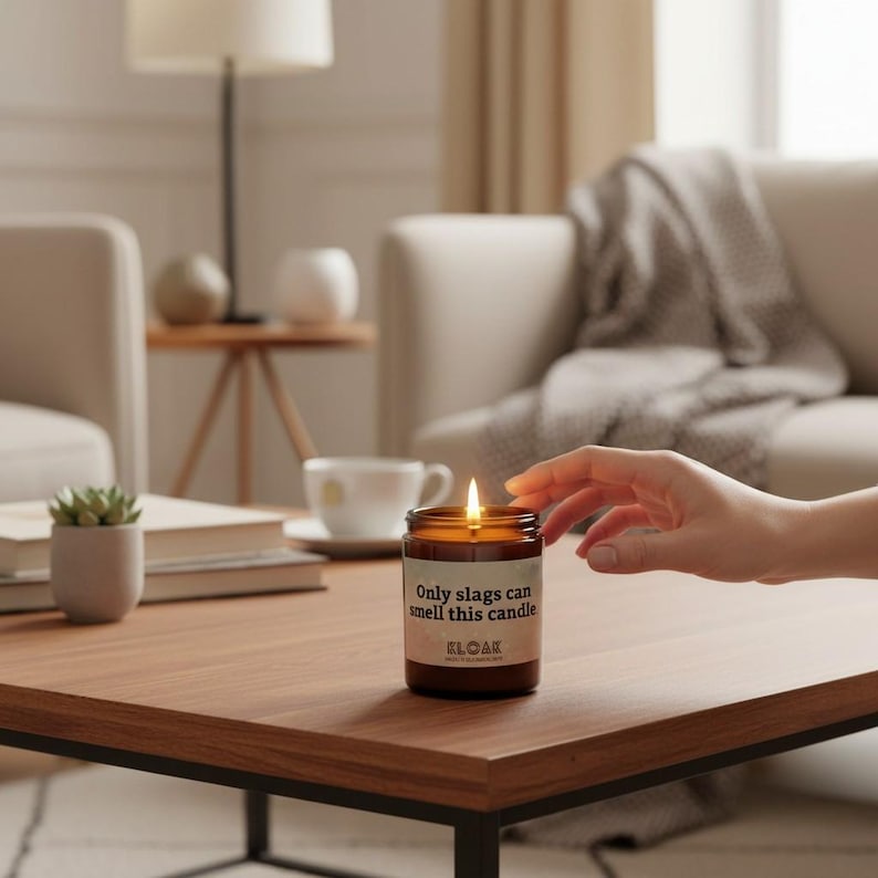 Puede incluir: Una vela encendida en un tarro de cristal marr&oacute;n con el texto "Only slags can smell this candle" y la marca "KLOAK" en una etiqueta. La vela est&aacute; sobre una mesa de centro de madera. Una mano se extiende hacia la vela. El fondo incluye un sof&aacute;, una l&aacute;mpara y otros elementos decorativos.