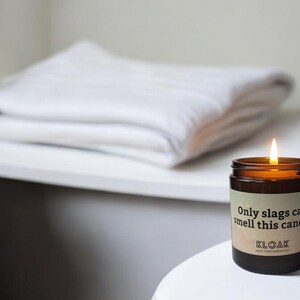 Puede incluir: Una vela de vidrio marr&oacute;n con una etiqueta blanca que dice "Only slags can smell this candle." La vela est&aacute; encendida y la llama es visible. La vela est&aacute; sobre una superficie blanca.