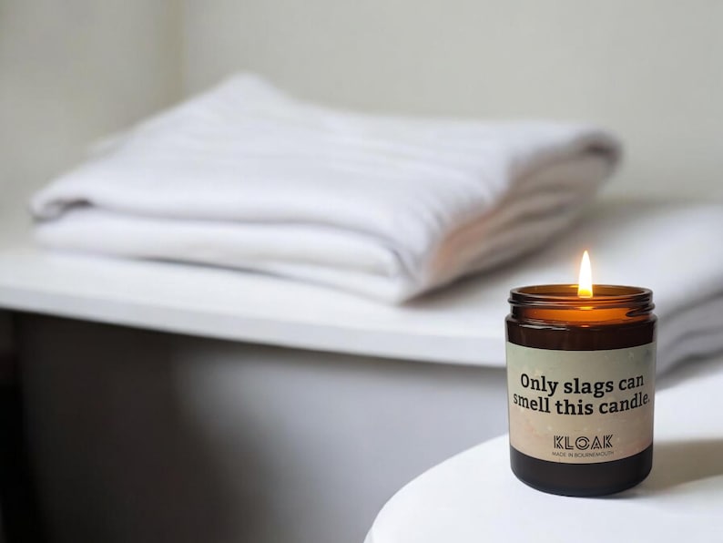 Puede incluir: Una vela de vidrio marr&oacute;n con una etiqueta blanca que dice "Only slags can smell this candle." La vela est&aacute; encendida y la llama es visible. La vela est&aacute; sobre una superficie blanca.