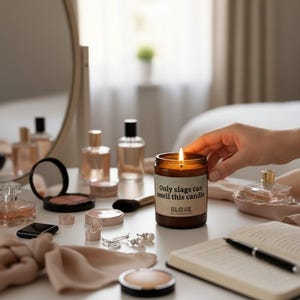Puede incluir: Una vela marr&oacute;n encendida con el texto "Only slags can smell this candle". La vela est&aacute; rodeada de frascos de perfume, maquillaje y un cuaderno sobre una mesa blanca.