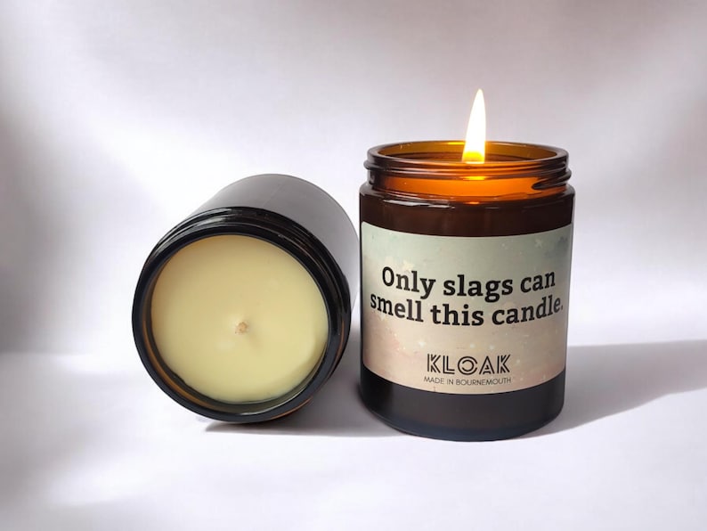 Puede incluir: Dos velas en frasco de vidrio &aacute;mbar con cera color crema. Una vela est&aacute; encendida con una llama visible. La etiqueta presenta un fondo degradado con estrellas y el texto: Only slags can smell this candle. KLOAK MADE IN BOURNEMOUTH.