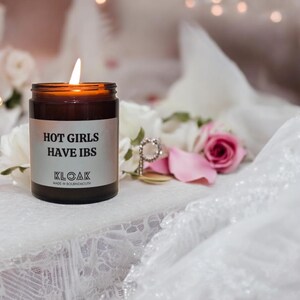 Pode incluir: Uma vela de vidro marrom com um r&oacute;tulo branco que diz "HOT GIRLS HAVE IBS" e "KLOAK MADE IN BOURNEMOUTH". A vela est&aacute; acesa e est&aacute; sobre uma toalha de renda branca com p&eacute;talas de rosa rosa.