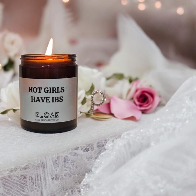 Pode incluir: Uma vela de vidro marrom com um r&oacute;tulo branco que diz "HOT GIRLS HAVE IBS" e "KLOAK MADE IN BOURNEMOUTH". A vela est&aacute; acesa e est&aacute; sobre uma toalha de renda branca com p&eacute;talas de rosa rosa.
