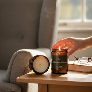 May include: A lit candle in a brown glass jar with a black lid sits on a wooden table. The candle has a label that reads "I love chicken wine" with a rooster graphic. A second candle is visible, along with a book and glasses.