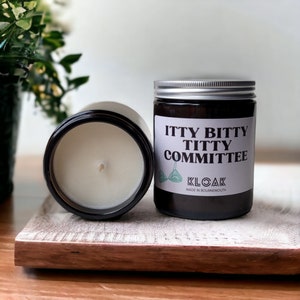 Op de afbeelding: Twee bruine glazen potkaarsen met zilveren deksels. Eén kaars ligt op zijn kant en onthult de witte was en lont. De andere kaars heeft een label met "ITTY BITTY TITTY COMMITTEE" met een bh-afbeelding en "KLOAK MADE IN BOURNEMOUTH".