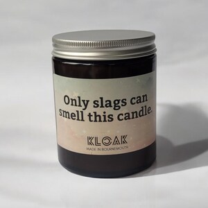 Puede incluir: Una vela en frasco de vidrio marr&oacute;n oscuro con tapa de rosca plateada. La etiqueta presenta un fondo degradado en tonos pastel con el texto: Only slags can smell this candle. KLOAK MADE IN BOURNEMOUTH impreso en negro en la parte inferior.