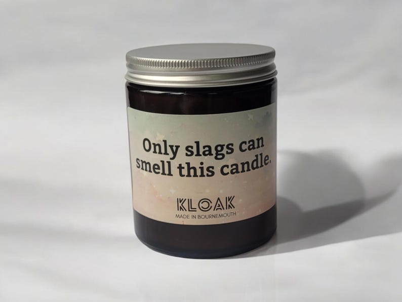 Puede incluir: Una vela en frasco de vidrio marr&oacute;n oscuro con tapa de rosca plateada. La etiqueta presenta un fondo degradado en tonos pastel con el texto: Only slags can smell this candle. KLOAK MADE IN BOURNEMOUTH impreso en negro en la parte inferior.