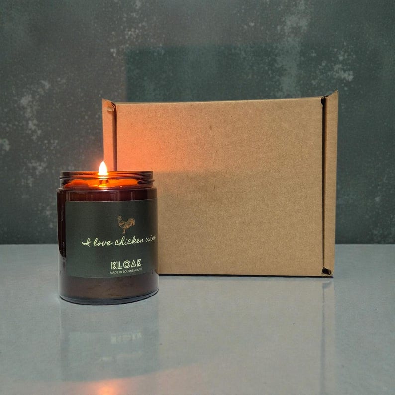 May include: A lit, brown glass jar candle with a label that reads "I love chicken wings" and a rooster graphic. A brown cardboard box is behind the candle. The candle is on a reflective surface.