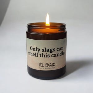 Puede incluir: Un tarro de vela de vidrio marr&oacute;n encendido con una etiqueta que dice "Only slags can smell this candle". La vela tiene una llama y la marca "KLOAK" es visible. La vela est&aacute; hecha en Bournemouth.