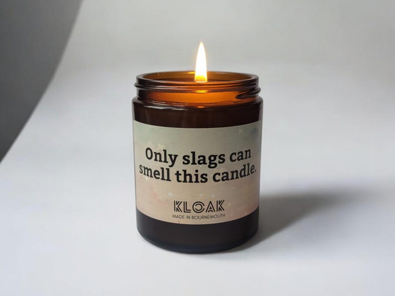 Puede incluir: Un tarro de vela de vidrio marr&oacute;n encendido con una etiqueta que dice "Only slags can smell this candle". La vela tiene una llama y la marca "KLOAK" es visible. La vela est&aacute; hecha en Bournemouth.