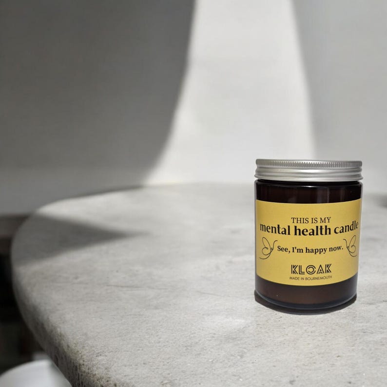 May include: A brown glass jar candle with a silver lid. The label reads "THIS IS MY mental health candle" with the text "See, I'm happy now." and the brand name "KLOAK". The candle is on a light grey surface.