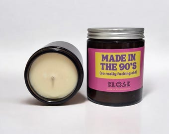 Op de afbeelding: Een zwarte glazen kaars pot met een zilveren deksel. De kaars is wit en heeft een houten lont. De pot heeft een roze label met de tekst "MADE IN THE 90's (so really fucking old)" en de tekst "KLOAK MADE IN BOURNEMOUTH".