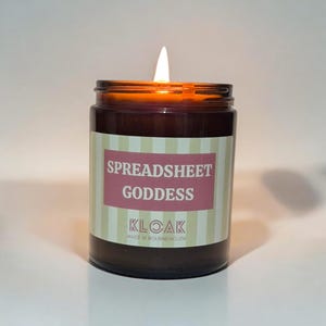 May include: A burning candle in a brown glass jar. The label features the text "SPREADSHEET GODDESS" in white on a pink background, with a striped design. The brand name "KLOAK" is also visible.