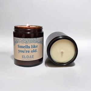 Könnte beinhalten: Ein braunes Glasgefäß mit einer Kerze und einem weißen Etikett mit der Aufschrift "Smells like you're old." Die Kerze ist angezündet und das Gefäß steht auf einer weißen Oberfläche. Die Kerze wird von KLOAK hergestellt und in Bournemouth hergestellt.
