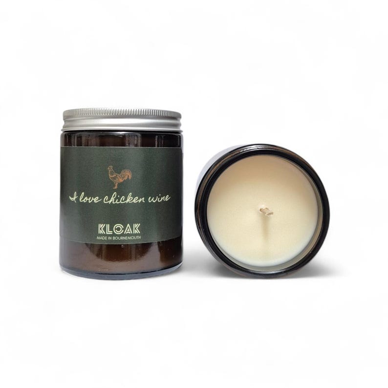 May include: Two brown glass jar candles with silver lids. One candle is upright, the other is on its side. The label on the upright candle reads "I love chicken wine" with a rooster illustration. The brand name "KLOAK" is also visible.