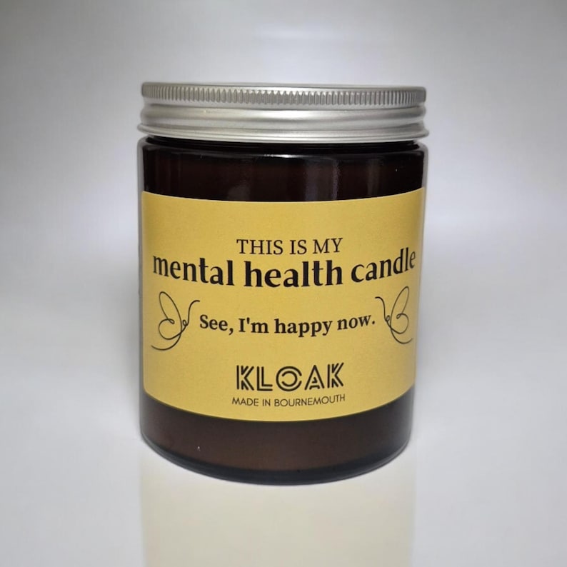 May include: A brown glass jar with a silver lid, labelled "THIS IS MY mental health candle." The label also includes the text "See, I'm happy now." and the brand name "KLOAK, MADE IN BOURNEMOUTH."