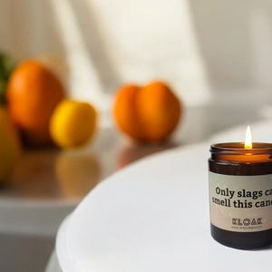 Puede incluir: Una vela encendida en un tarro de cristal marr&oacute;n con el texto "Only slags can smell this candle" en una etiqueta blanca. La vela est&aacute; sobre una superficie blanca con naranjas y limones borrosos en el fondo.