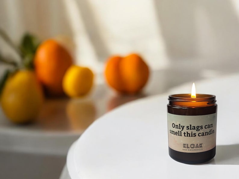 Puede incluir: Una vela encendida en un tarro de cristal marr&oacute;n con el texto "Only slags can smell this candle" en una etiqueta blanca. La vela est&aacute; sobre una superficie blanca con naranjas y limones borrosos en el fondo.