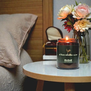 May include: A lit candle in a glass jar with a dark green label that reads "I love chicken wine" and features a rooster illustration. The candle sits on a white table next to a vase of flowers.