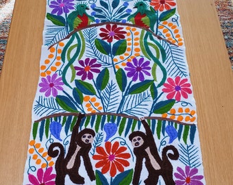 May include: A white table runner with colorful embroidered flowers, birds, and monkeys. The design features a repeating pattern of green leaves, orange dots, and blue vines.