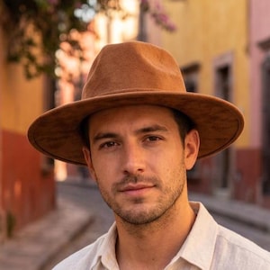 Könnte beinhalten: Ein breitkrempiger Fedora-Hut in einfarbigem Hellbraun mit strukturierter Krone und passendem, farblich abgestimmtem Band. Der Hut hat eine glatte, filzartige Textur und eine steife Krempe, die den Kopf umschließt.
