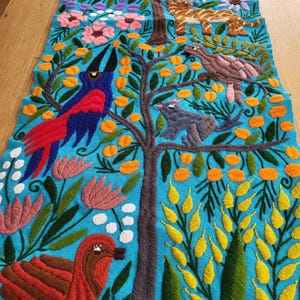 Könnte beinhalten: Ein hellblauer Textilläufer mit aufwendigen Stickereien eines braunen Baumes, eines roten Vogels, eines blauen Papageis, eines grauen Eichhörnchens, einer beigen Katze sowie diverser bunter Blumen und gelbem Blattwerk. Das Design zeigt Details.