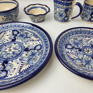 Könnte beinhalten: Eine Sammlung von blau-weißen Keramikgeschirr, darunter Schalen, Becher und ovale Platten. Die Stücke weisen ein Blumendesign mit komplizierten Details und einem dunkelblauen Rand auf. Das Set ist auf einer weißen Oberfläche angeordnet.