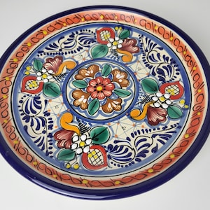 Könnte beinhalten: Eine handbemalte Keramikplatte mit einem lebendigen Blumendesign. Der Teller hat einen dunkelblauen Rand, eine hellblaue Mitte und einen Rand mit orangefarbenen und roten Akzenten. Das Blumenmuster enthält rote, orangefarbene, grüne und weiße Details.