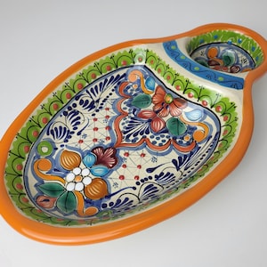 Pode incluir: Uma travessa de servir em cerâmica pintada à mão com um design floral vibrante. A travessa apresenta uma borda laranja, uma seção central com padrões intrincados em azul, verde e laranja e uma pequena tigela anexada. A forma é oval com uma alça.