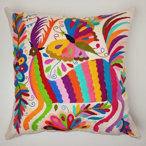 Puede incluir: Una almohada bordada colorida con un gallo con una mariposa en la espalda. La almohada está hecha de tela blanca y tiene una variedad de colores brillantes, incluyendo rojo, naranja, amarillo, verde, azul, morado y rosa.